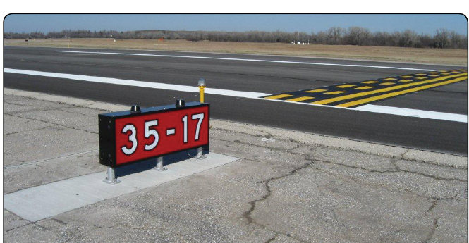 A ground-level photograph of a low red sign panel mounted on three short legs on a concrete pad. The panel reads 35-17 in white characters outlined in black. Beyond the pad the pavement runs out to a dark surface bounded by white stripes. In the right half of the frame a holding position marking crosses that pavement diagonally, drawn as four yellow lines outlined in black, the two nearer lines solid and the two farther lines dashed. Trees and open ground lie along the horizon under a clear sky.