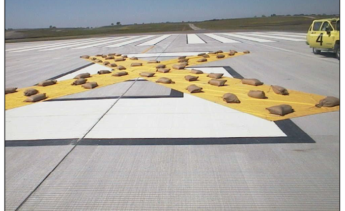 A large yellow vinyl cross laid flat across the white threshold stripes of a concrete runway, weighted down by rows of sandbags. A yellow airfield vehicle stands at the right edge of the frame and open country lies beyond the runway end.