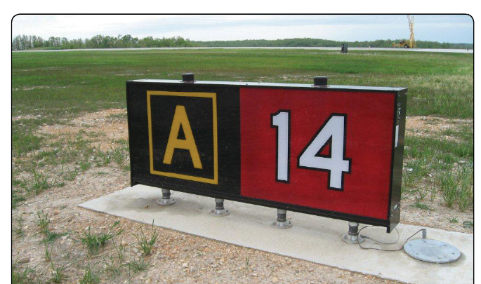 A ground-level photograph of a single sign panel assembly standing on a concrete pad in the grass beside an airfield, with a runway and open country behind it. The left half of the panel is black and carries a yellow letter A inside a yellow border. The right half is red and carries the number 14 in white with a black outline.