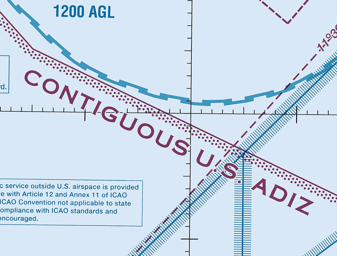 Sectional chart excerpt over the Pacific Ocean on a pale blue tint with no land in the frame. A band of magenta dots runs from the upper left to the lower right beside a thin solid magenta line, with the words CONTIGUOUS U.S. ADIZ lettered in large magenta capitals along it. Above the band a heavy blue line made of rectangular blocks curves across the frame, labeled 1200 AGL, and at the lower right two blue lines each carrying a band of long thin ticks cross one another.