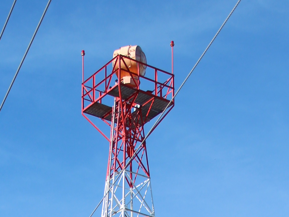 Looking up from the ground at the top of a red and white lattice tower against a clear blue sky. A rotating beacon unit with a pale housing and a tinted lens face stands on a red railed platform at the top of the tower. A small red obstruction light is mounted on a short mast at each of two platform corners. Three cables cross the frame diagonally.