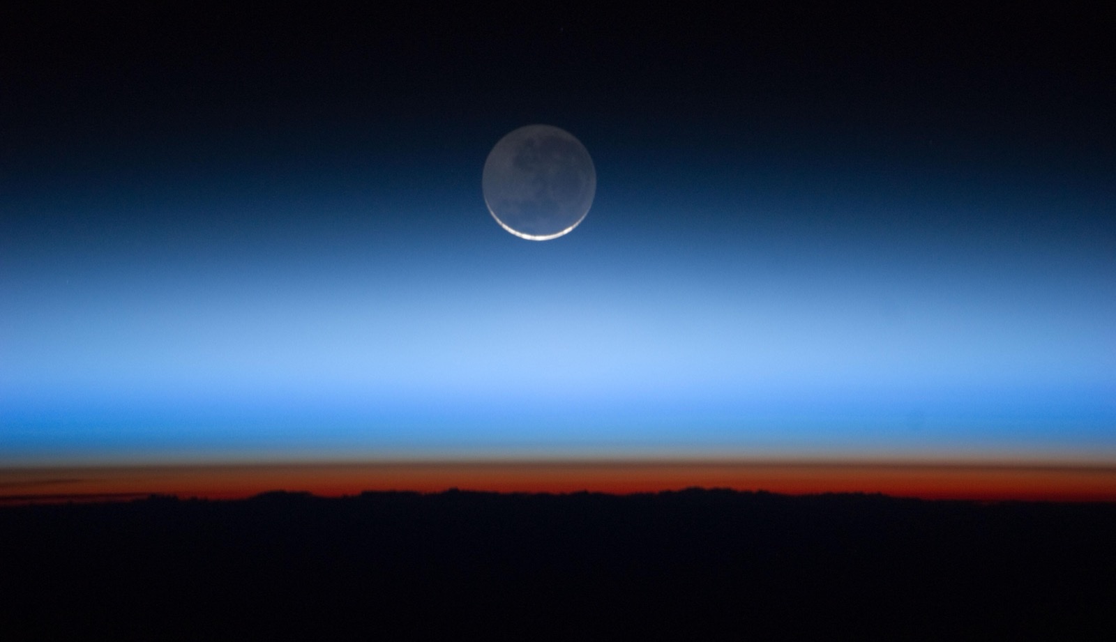 The edge of the Earth's atmosphere photographed from orbit against the black of space. A dark silhouetted cloud deck fills the bottom of the frame. Immediately above it lies a narrow red-orange band, then a paler grey-white band, then a broad bright blue band that fades upward into black. A crescent-lit Moon hangs in the black above the blue.