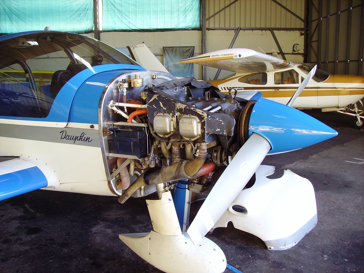 A four-cylinder horizontally opposed piston engine mounted in an airplane nose with the cowling removed, showing two cylinders on each side, the blue spinner, and one propeller blade, in a hangar