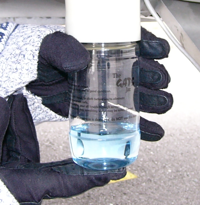 A gloved hand holding a clear plastic fuel tester filled with visibly blue fuel drawn from an aircraft sump, held up against a neutral background to check its color and clarity