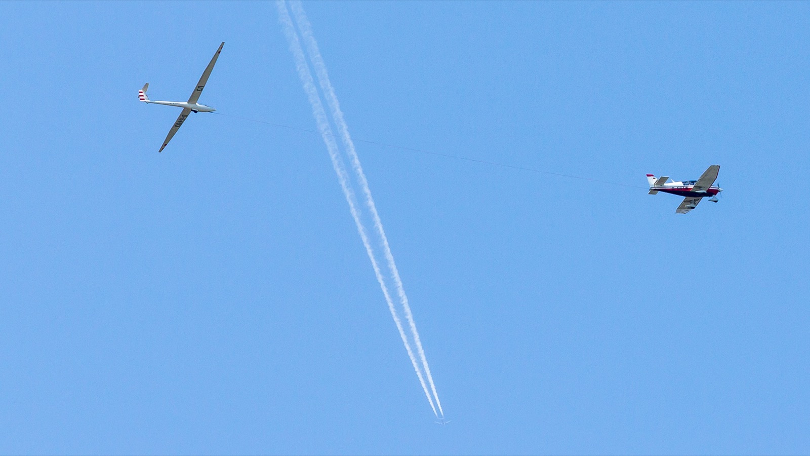 A single-engine Robin towplane climbing with a sailplane behind it on a taut towline, both aircraft banking gently over green river valley terrain.