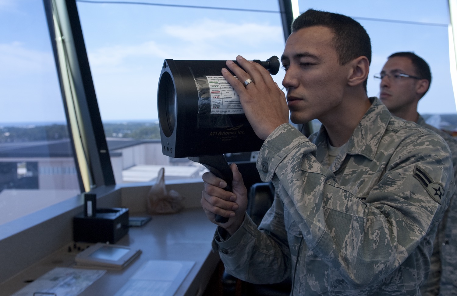 Inside a control tower cab, an air traffic controller in uniform holds a black signal light gun to his eye with both hands and aims it out through the sloping tower window. The device has a wide cylindrical barrel with a lens at the front, a pistol grip below, and a small eyepiece at the back. A printed card listing the signals and their meanings is taped to the side of the barrel. A second controller stands behind him, and the airfield and a low building are visible through the glass.