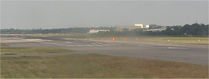 A hazy view across an airport in flat light. A runway runs from left to right across the lower half of the frame. Beyond its far edge, four evenly spaced single light units in one row all show red. Terminal buildings and trees stand on the horizon.