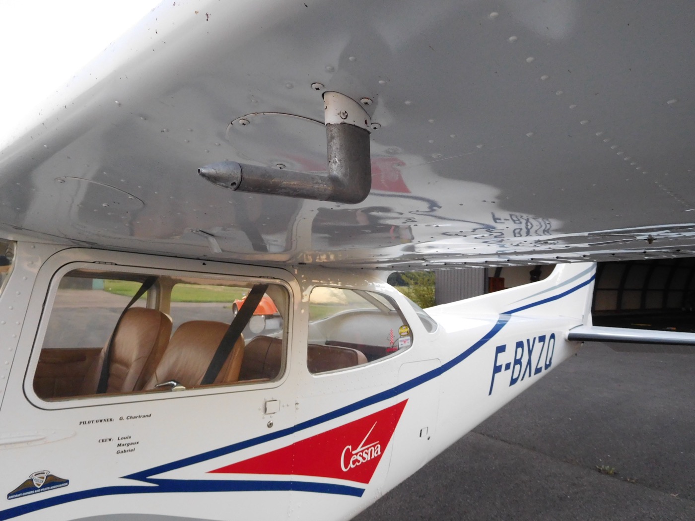 An L-shaped metal pitot tube mounted on a strut under the left wing of a white Cessna, its open front end facing forward into the airflow, with the cabin windows and registration below
