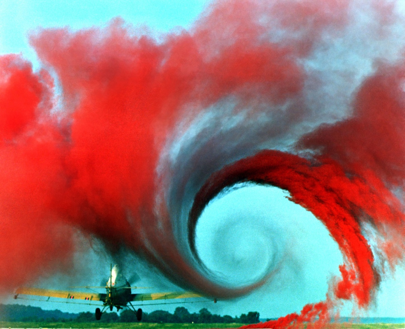 A crop duster flying low over a field with colored smoke drawn into a huge spiral behind it, making the rotation of the trailing wingtip vortex visible