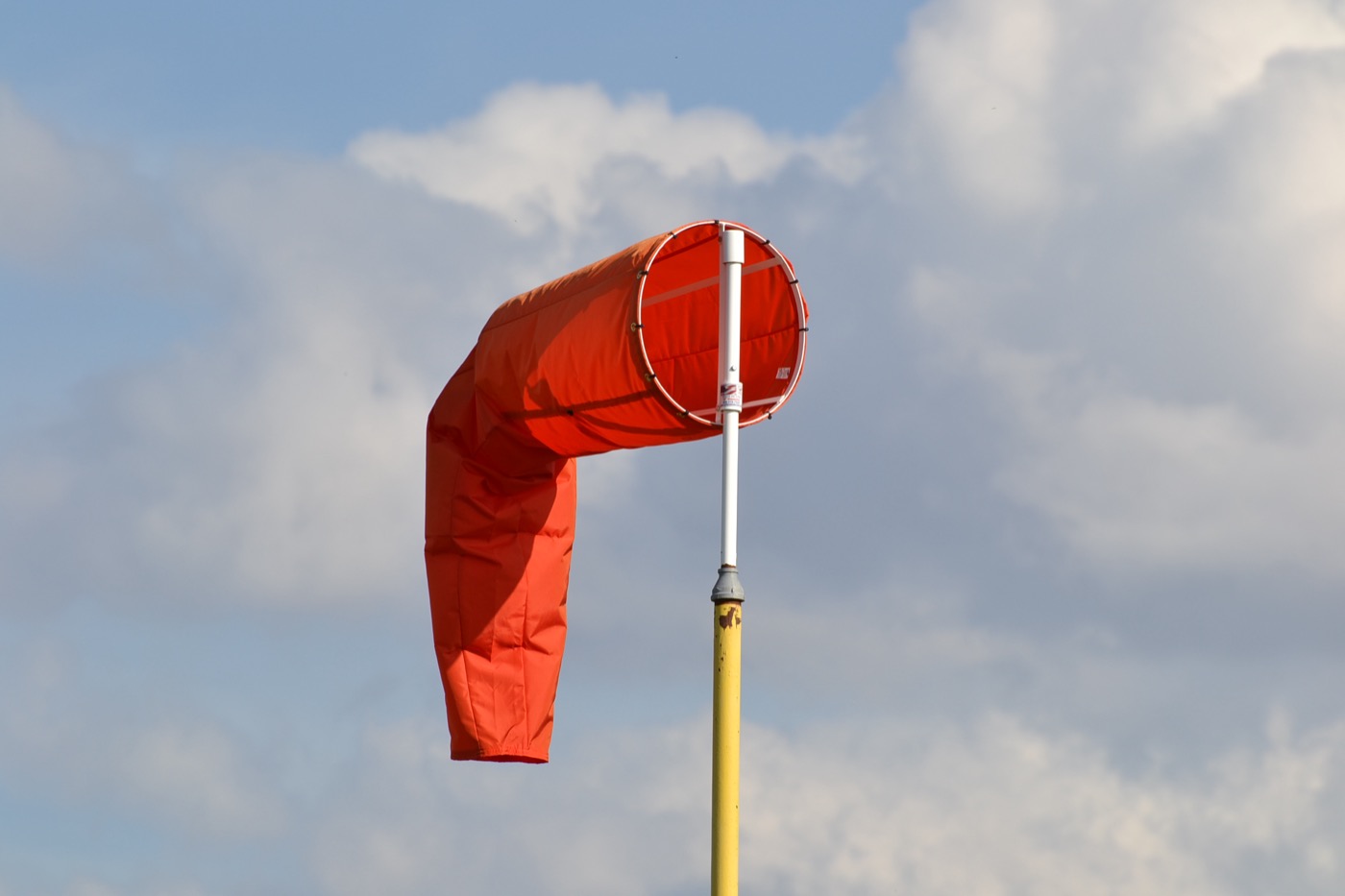 An orange wind cone on a yellow pole against a partly cloudy sky. The cone's large end is attached at the top of the pole and its narrow end trails away and downward, so the fabric is flying at roughly 45 degrees below horizontal rather than standing straight out.
